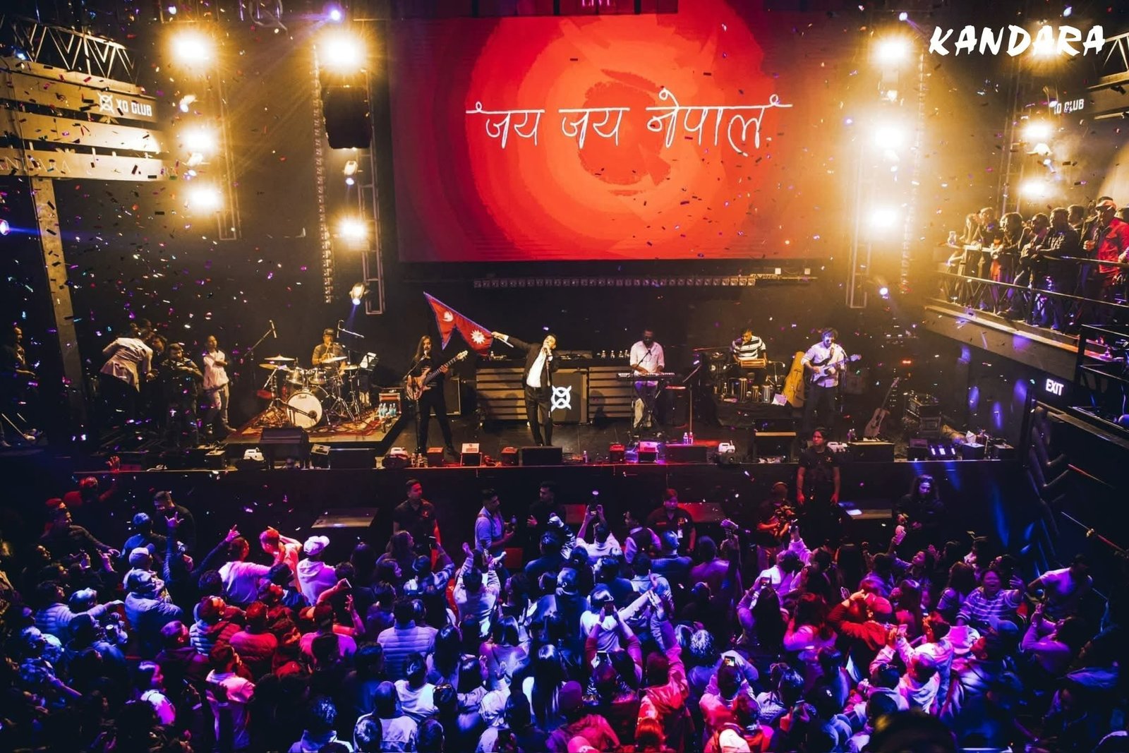 Kandara on stage under a red backdrop, a Nepali flag raised mid-song, confetti over a full standing crowd.